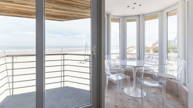 Un balcon avec vue sur la plage, des chaises en verre transparent et une table blanche.