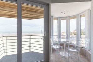 Un balcón con vista a la playa, sillas de vidrio transparente y una mesa blanca.