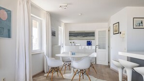 Une cuisine moderne avec des meubles blancs, une table de salle à manger ronde entourée de quatre chaises, un coin bar avec des tabourets et une grande fenêtre avec des rideaux.
