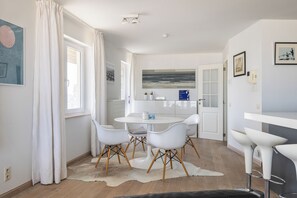 A modern kitchen with white cabinetry, a round dining table with four chairs, a bar area with stools, and a large window with curtains. - Malibu Beach 0201 + P* (Sint Idesbald)