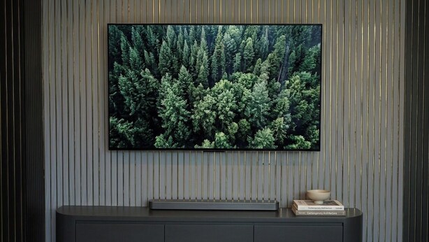 Un televisor de pantalla plana montado en la pared con una imagen de un bosque, una consola oscura debajo y un cuenco pequeño y un libro sobre la consola.