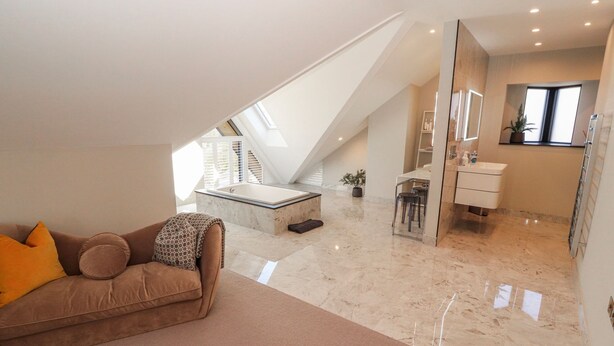 A modern loft bedroom with a sloped ceiling, a built-in bathtub, a sitting area with a sofa and cushions, and a bathroom with a vanity and mirror. - Midways (Windermere)