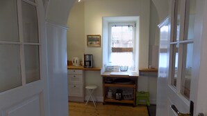 En smal hall leder in till ett kök med ett fönster, en kaffebryggare och en liten pall.