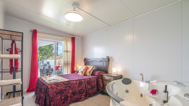 A bedroom with a bed, a nightstand, a lamp, a red curtain, a window, and a bathtub. - Apartment 'El Encanto' with Mountain View, Private Pool and Wi-Fi (Villarrasa)