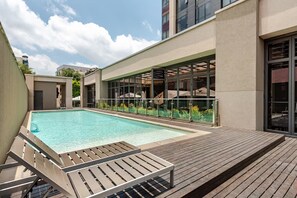 Ein Schwimmbad mit Holzterrasse, Glasgeländern und einem Gebäude mit großen Fenstern.