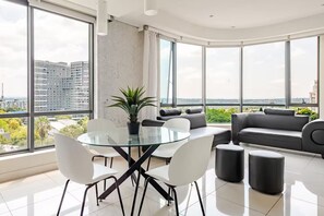A modern living room with a glass table, white chairs, a grey sofa, and a potted plant. - Sandton Skye | Chic Luxury Apartment (Sandton)