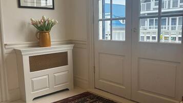 Una habitación con una puerta blanca, un cuadro en la pared, un florero con flores y una alfombra estampada.