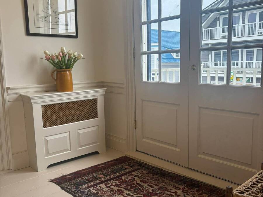 Una habitación con una puerta blanca, un cuadro en la pared, un florero con flores y una alfombra estampada.
