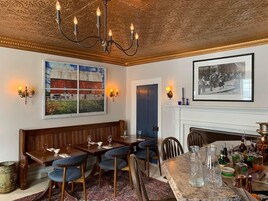 A dining area with a wooden bench, tables, and chairs, a chandelier, wall-mounted sconces, and a fireplace with a framed historical photo.