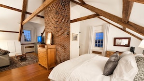Un dormitorio con chimenea de ladrillo, vigas de madera, cama con ropa de cama blanca, cómoda y escritorio con computadora.