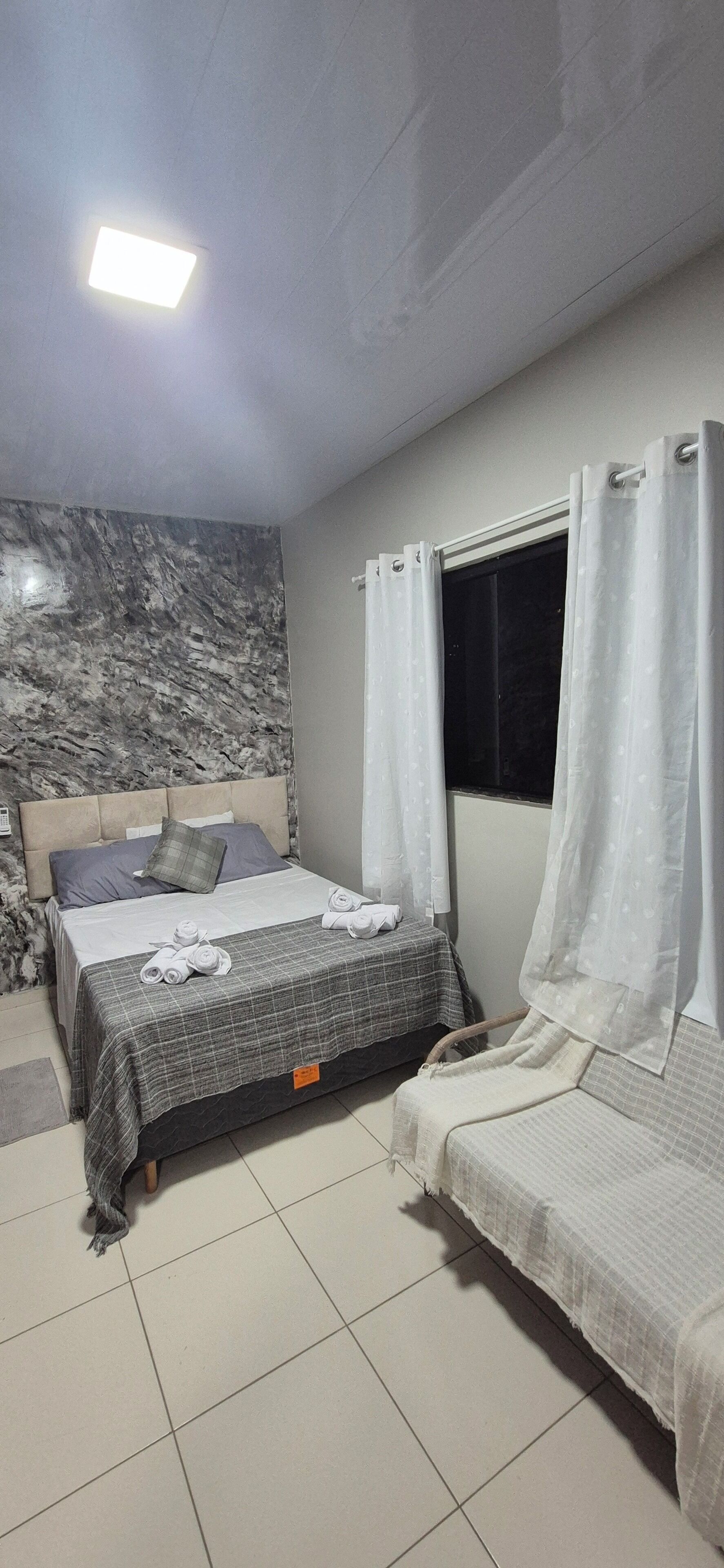 A bedroom with a bed, a smaller bed, a window with curtains, a marble wall, and a ceiling light.