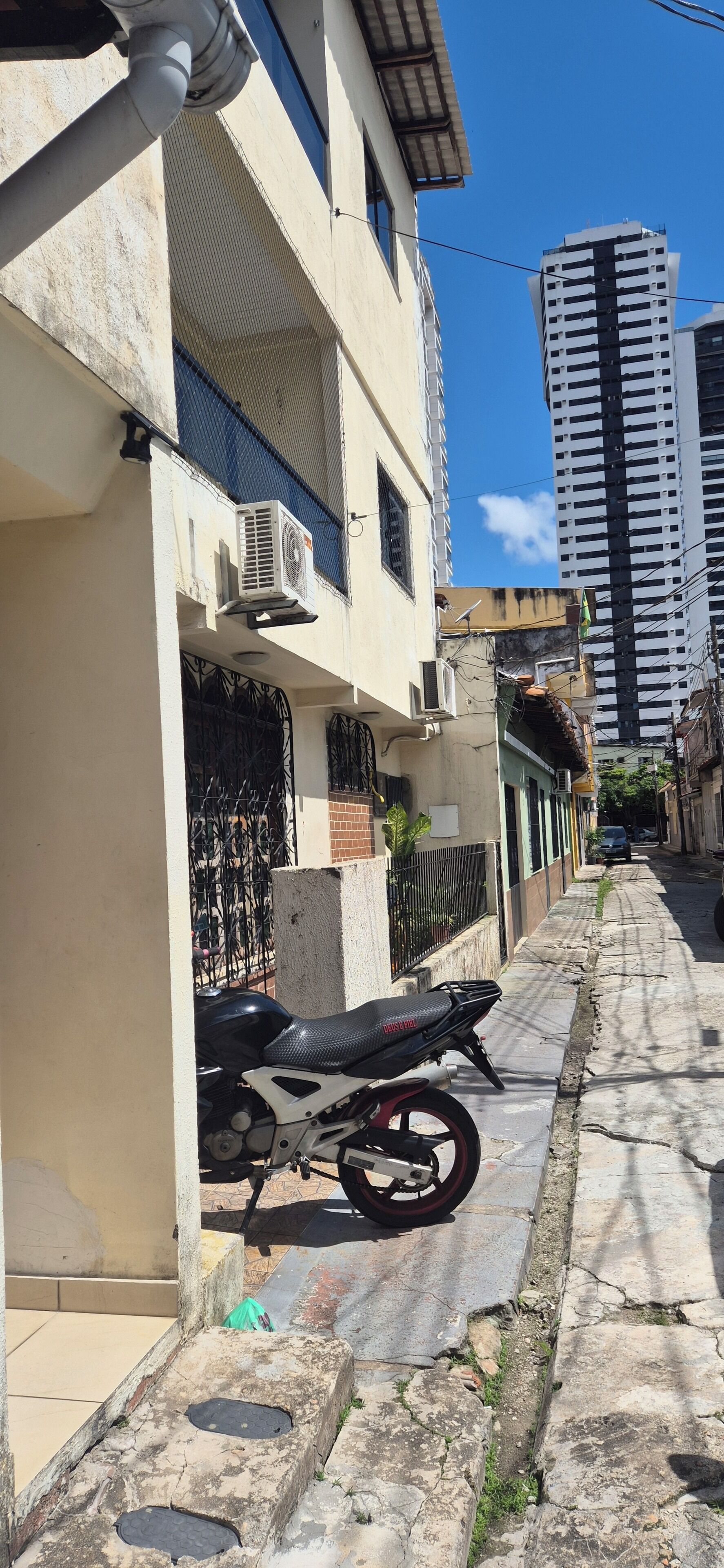 Un carrer urbà estret amb una moto aparcada al costat del carrer, un edifici amb un balcó i un gratacels alt al fons.