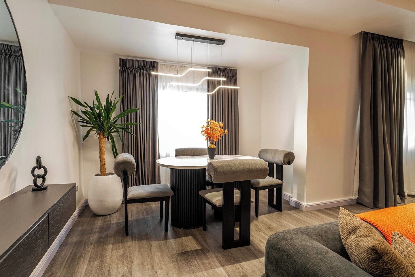 Una habitación de hotel moderna con zona de comedor, una planta grande en maceta, una mesa redonda y un sofá.