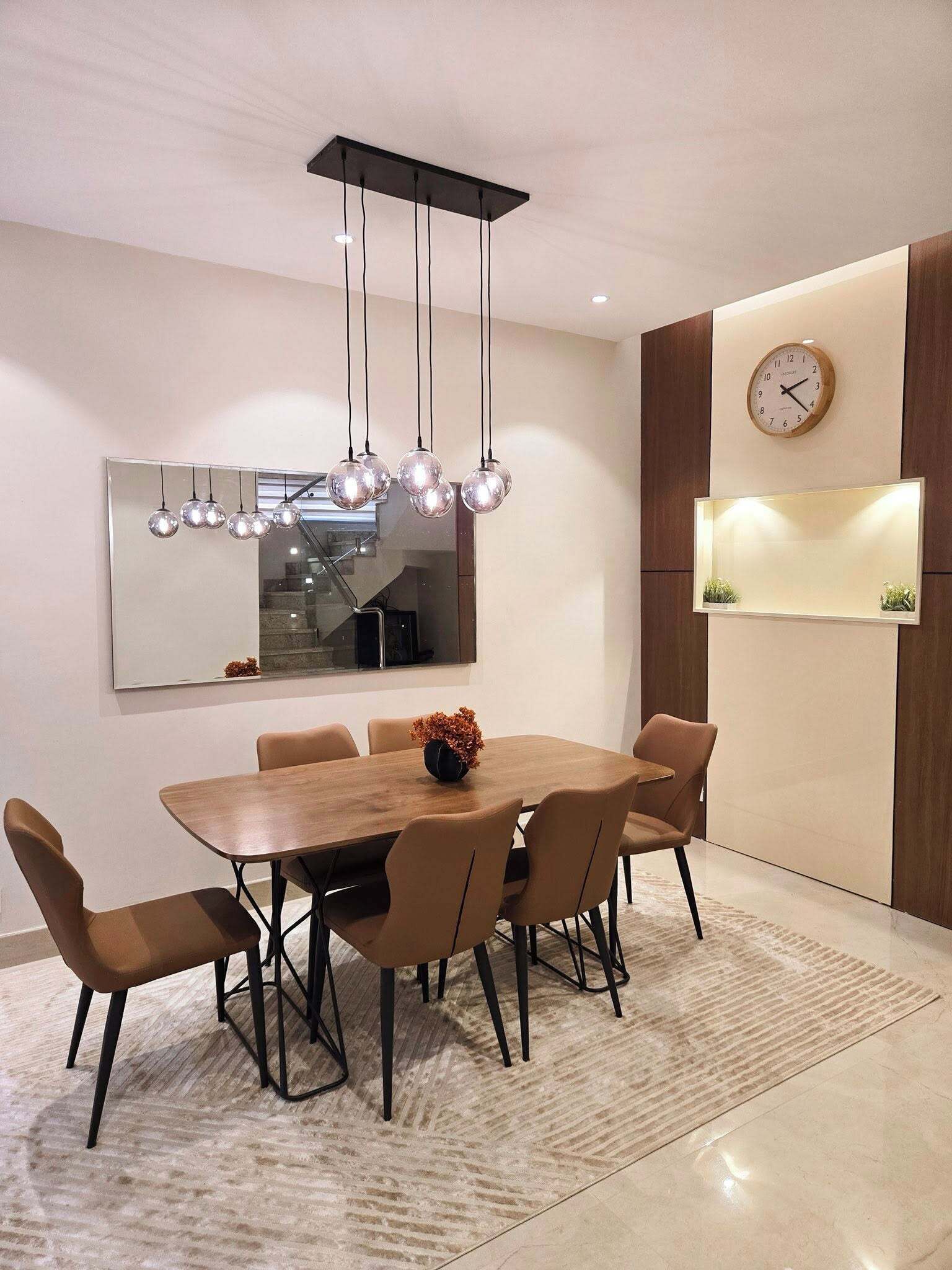A modern dining room with a wooden table, six chairs, a hanging spherical light fixture, a wall-mounted clock, and a mirror.