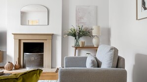 Ein Wohnzimmer mit Kamin, einer Couch, einem Spiegel und einer Vase mit Blumen.