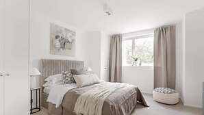 Een slaapkamer met een bed, nachtkastjes, een raam met gordijnen, een schilderij aan de muur en een klein, rond voetenbankje.