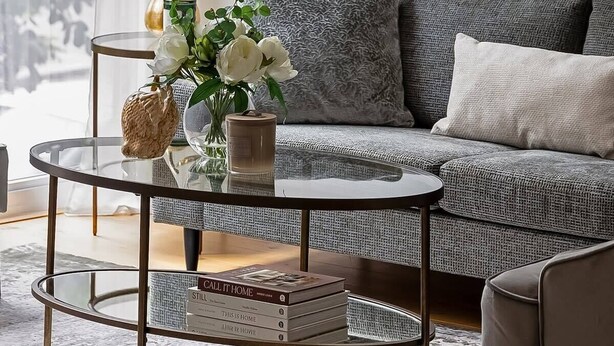 Een woonkamer met een grijze bank, een ronde salontafel met boeken, een vaas met bloemen en een schilderij aan de muur.