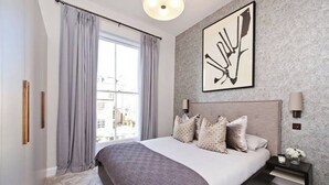Ein Schlafzimmer mit einem großen Bett, einem Fenster mit Vorhängen, einem Wandbild und einem Nachttisch mit Lampe.