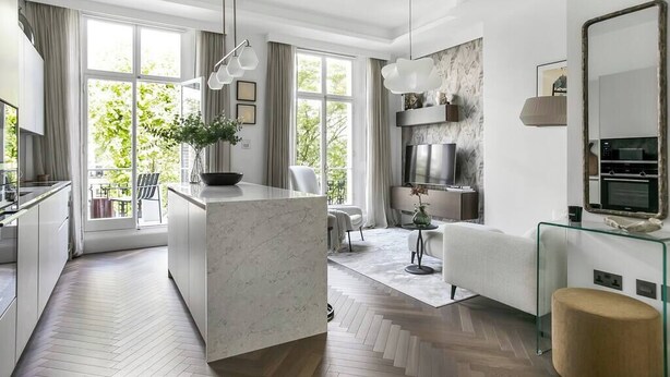 A modern kitchen with a marble island, a dining area with a glass table, and a living room with a sofa and TV.