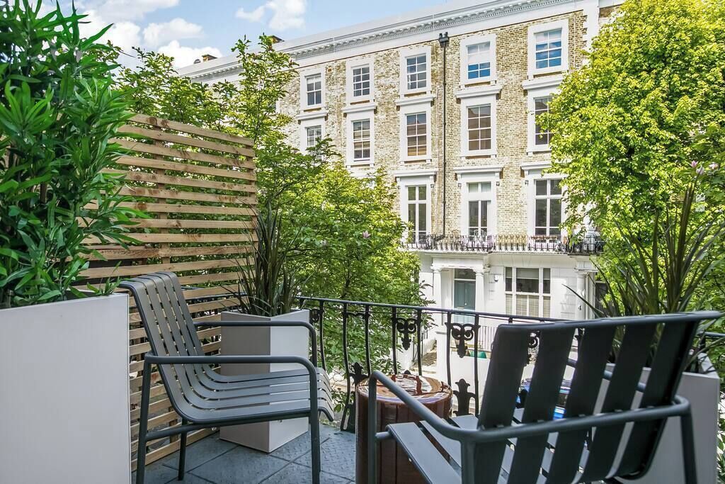 A balcony with outdoor seating, a view of a building with multiple windows, and some greenery.