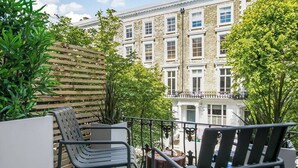 A balcony with outdoor seating, a view of a building with multiple windows, and some greenery.