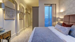 Ein Schlafzimmer mit einem großen Bett, einem Nachttisch, einem Schreibtisch, einem Balkon mit Vorhängen und Blick zum Himmel.
