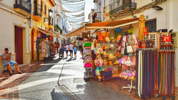 A narrow street with shops on both sides, a man sitting on a bench, and a variety of bags and hats displayed outside. - Sunny Studio in Nerja Center with Balcony close to Beach (Nerja)