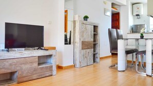 Een moderne woonkamer met een flatscreen televisie, een houten tv-meubel, een inbouwkast, een eethoek met een glazen tafel en stoelen, en een keuken met een boiler.
