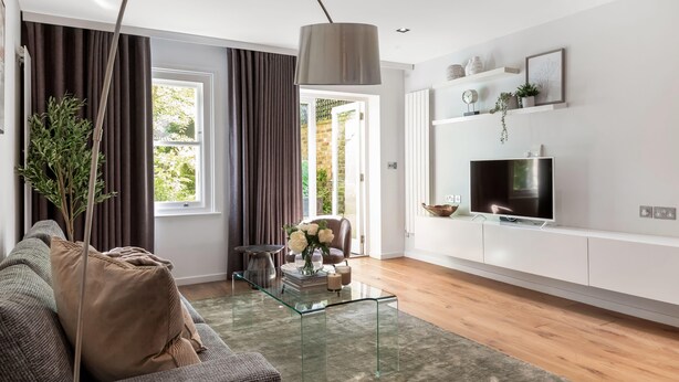 Ett modernt vardagsrum med en soffa, ett soffbord i glas, en tv på ett vitt skåp och ett stort fönster med gardiner.