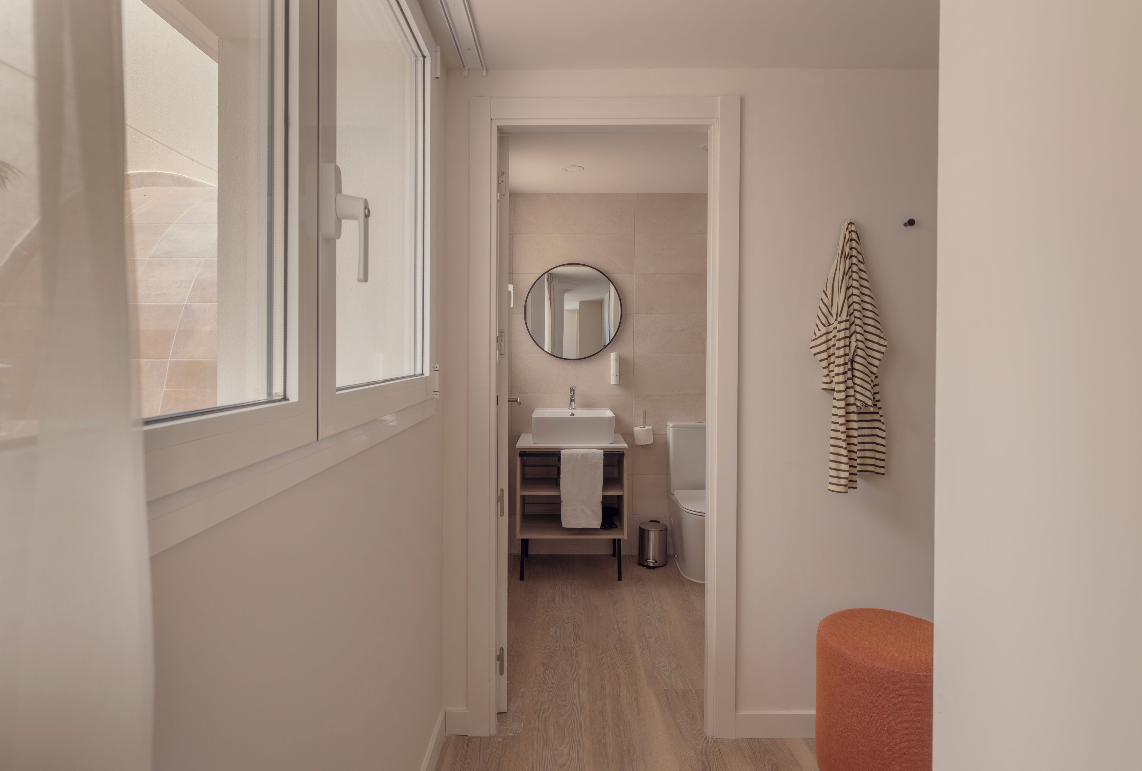 A narrow hallway leading to a bathroom with a window, a sink, a mirror, and a towel rack.
