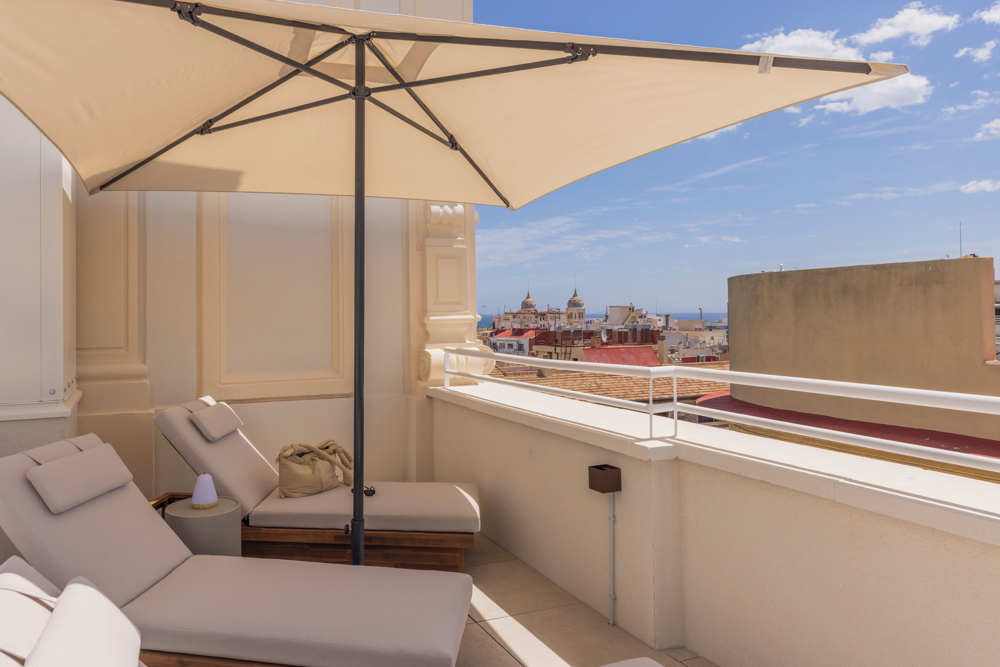 A balcony with a beige umbrella, a lounge chair, and a view of buildings.