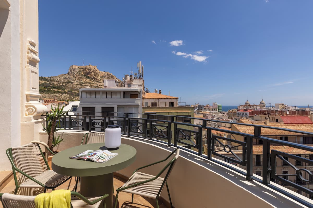 A balcony with a table, chairs, and a magazine, overlooking a cityscape with buildings and a hill.