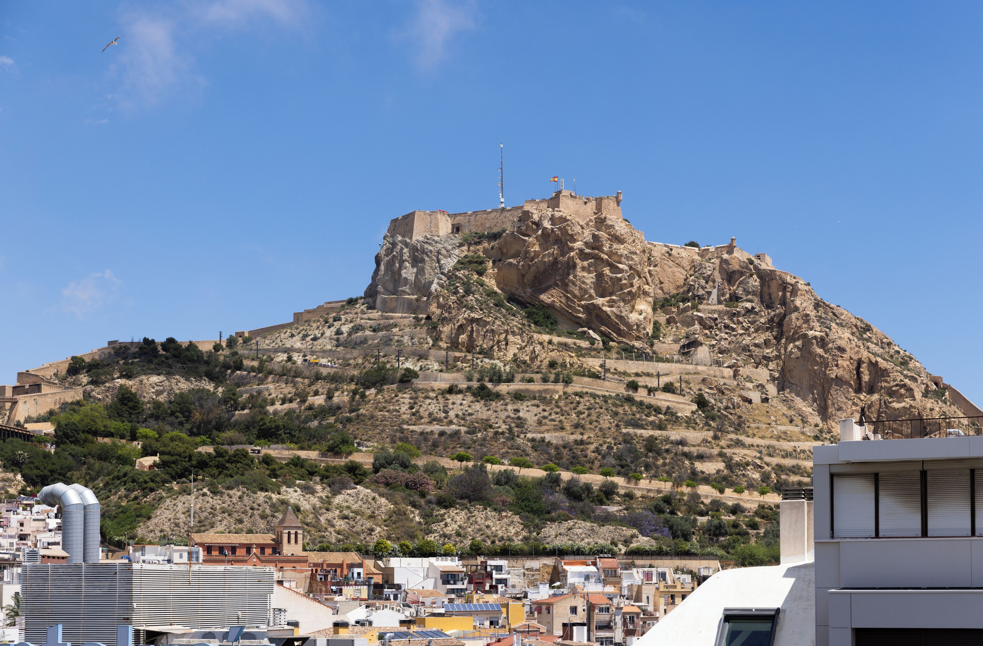 Un turó rocos on hi ha un edifici, envoltat d'una població amb diversos edificis.