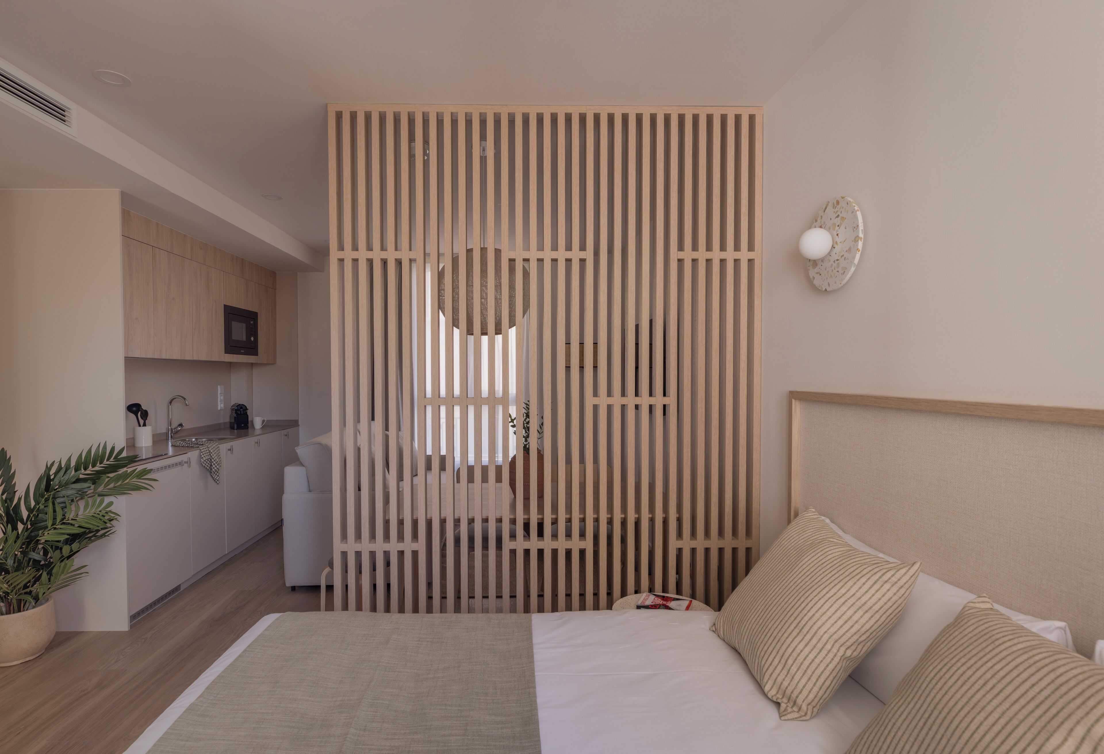 Een moderne hotelkamer met een houten scheidingswand, een bed met kussens, een nachtkastje, een potplant en uitzicht op de keuken.