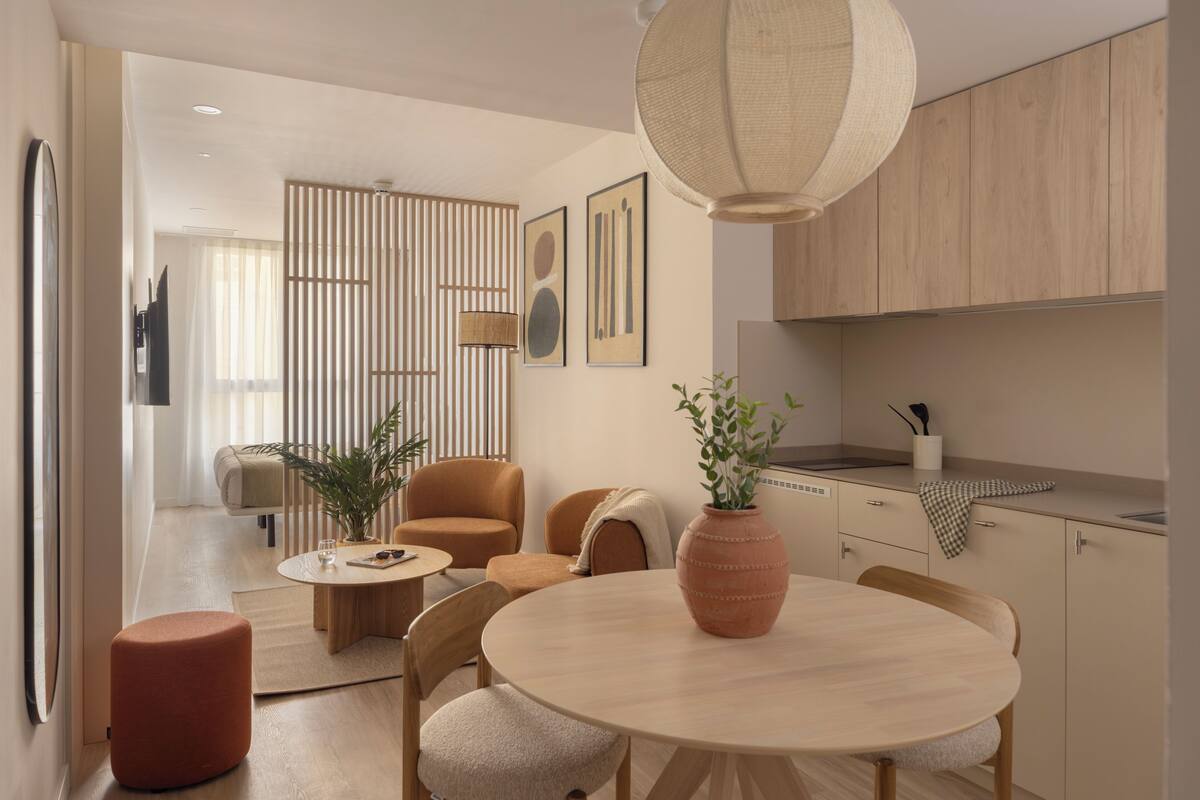 A modern interior with a round dining table, two chairs, a potted plant, and a kitchen area with wooden cabinets.