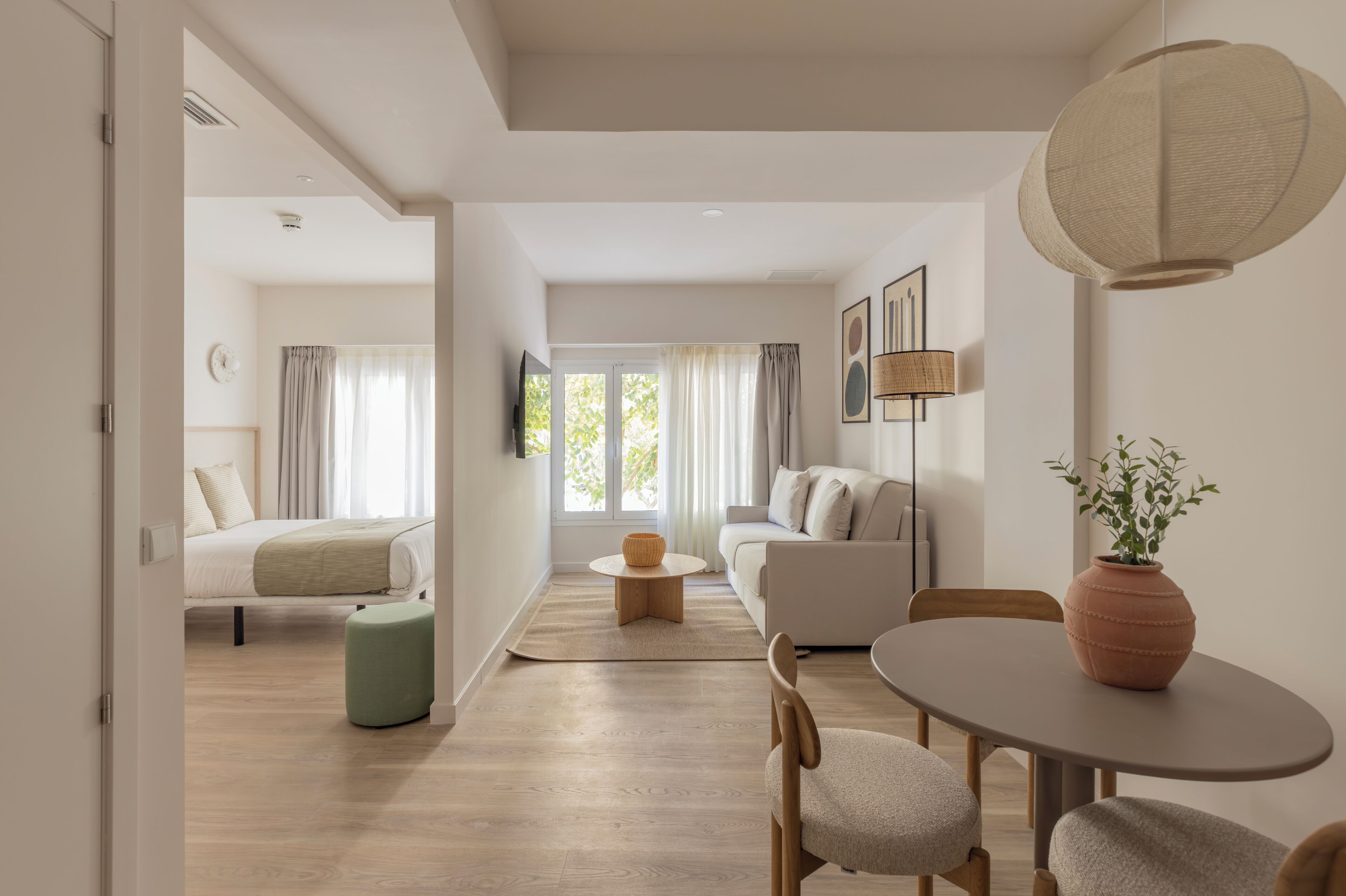 A modern hotel room with a bed, sofa, dining table, and a view of the outdoors.