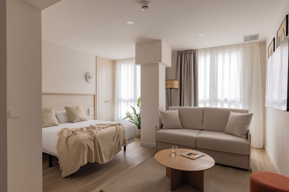 A modern hotel room with a bed, sofa, round coffee table, and a potted plant.