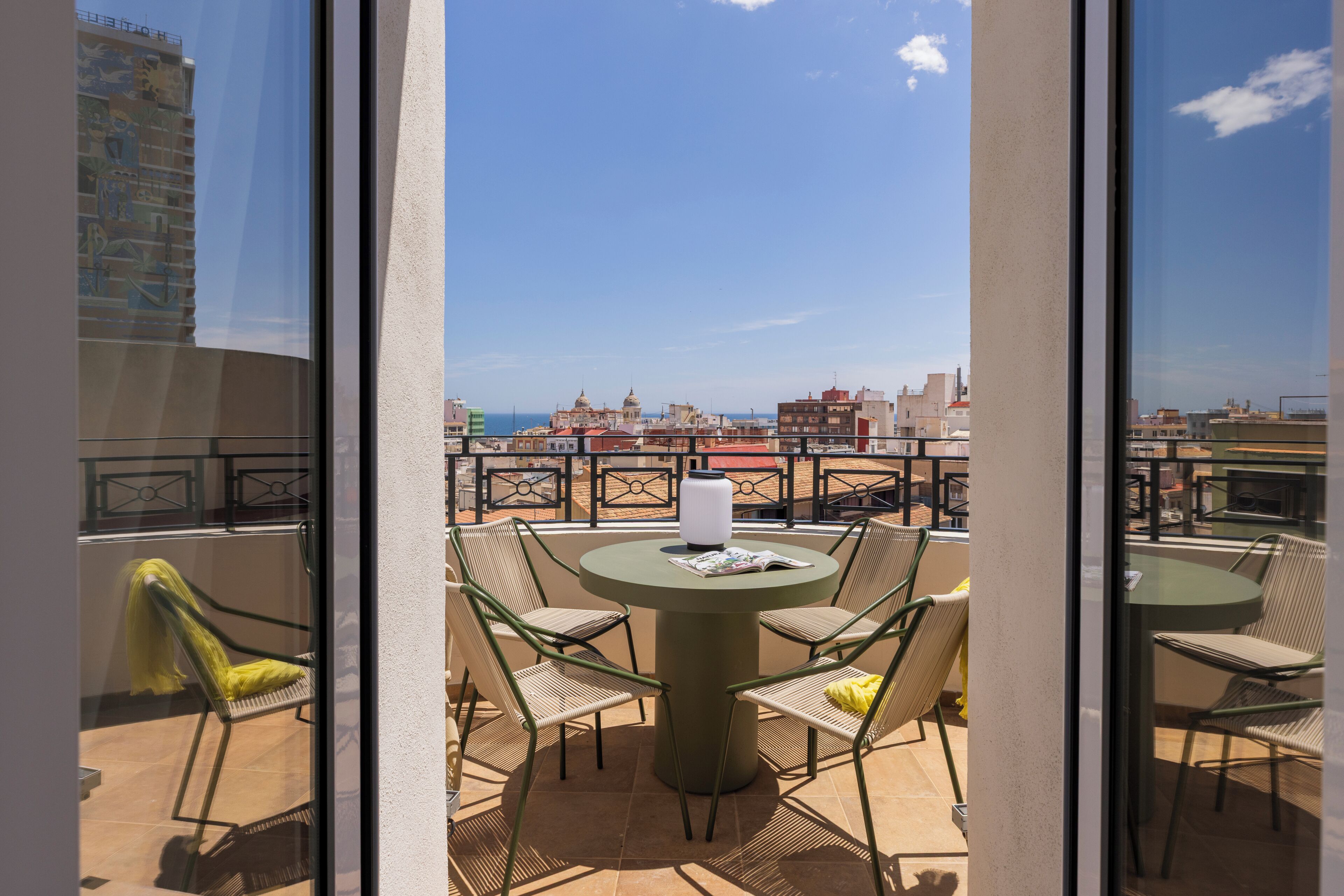 Un balcón con una mesa redonda y sillas, con vistas a un paisaje urbano que incluye edificios y un cielo despejado.