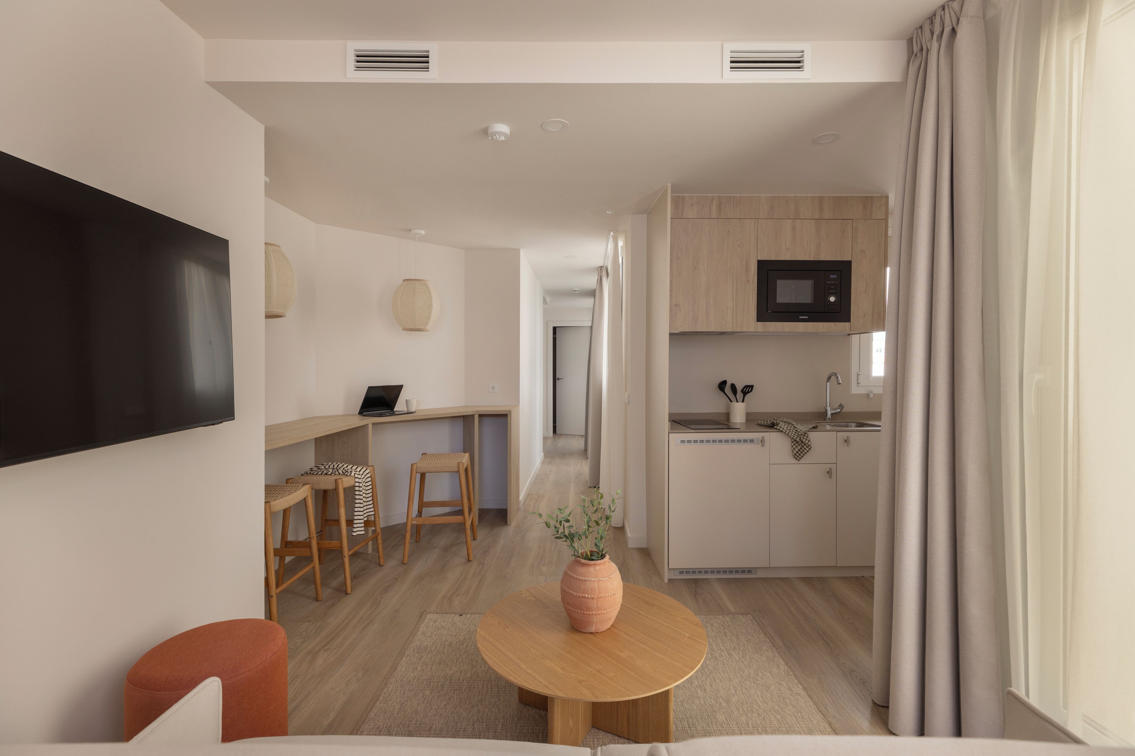 Una sala moderna con un televisor de pantalla plana, una mesa redonda de madera con un florero, un sofá y una cocina con armarios de madera y un microondas.