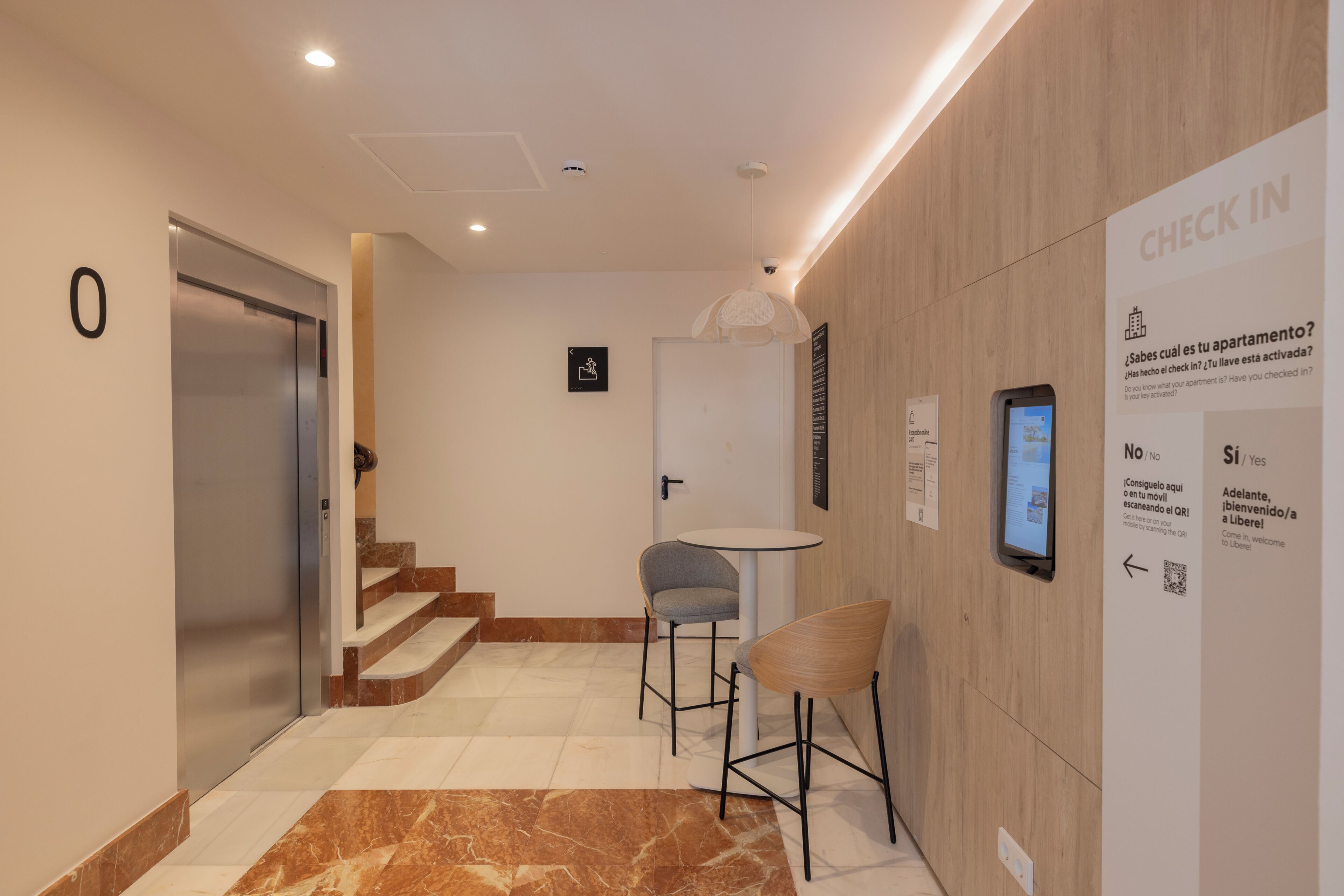 A modern hotel lobby with a check-in kiosk, a seating area, and a staircase leading to another floor.