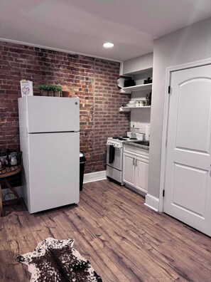 Cocina compacta con pared de ladrillo, armarios blancos, un refrigerador, un horno y una puerta.