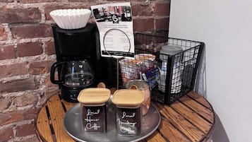 Una estación de café con una cafetera, dos tarros etiquetados 'Local Coffee', una bandeja con una taza de café y un cartel publicando cafeterías cercanas.