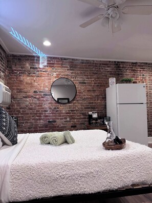 Un dormitorio con pared de ladrillo, un espejo redondo, una cama con colcha blanca texturizada, un ventilador de techo y un refrigerador blanco.