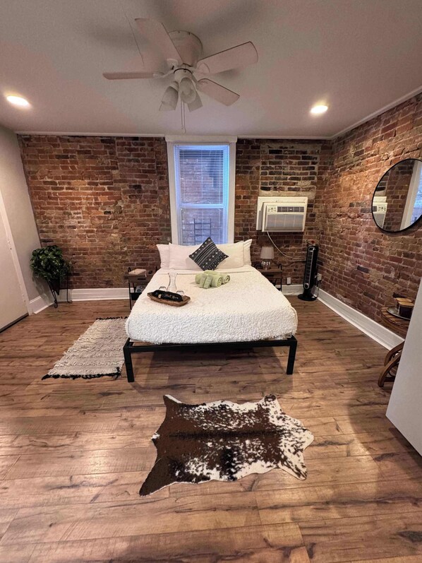 Un dormitorio con pared de ladrillo, cama con ropa de cama blanca, ventilador de techo, ventana con persianas y piso de madera.