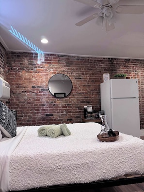 Un dormitorio con pared de ladrillo, un espejo redondo, una cama con colcha blanca texturizada, un ventilador de techo y un refrigerador blanco.