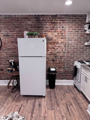 Una cocina con un refrigerador blanco, un cubo de basura, una cafetera y una pared de ladrillo.