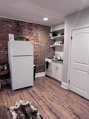Cocina compacta con pared de ladrillo, armarios blancos, un refrigerador, un horno y una puerta.