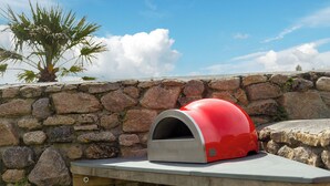 A stone wall with a red and silver outdoor oven on a wooden ledge. - Scilly Stack (Penzance)