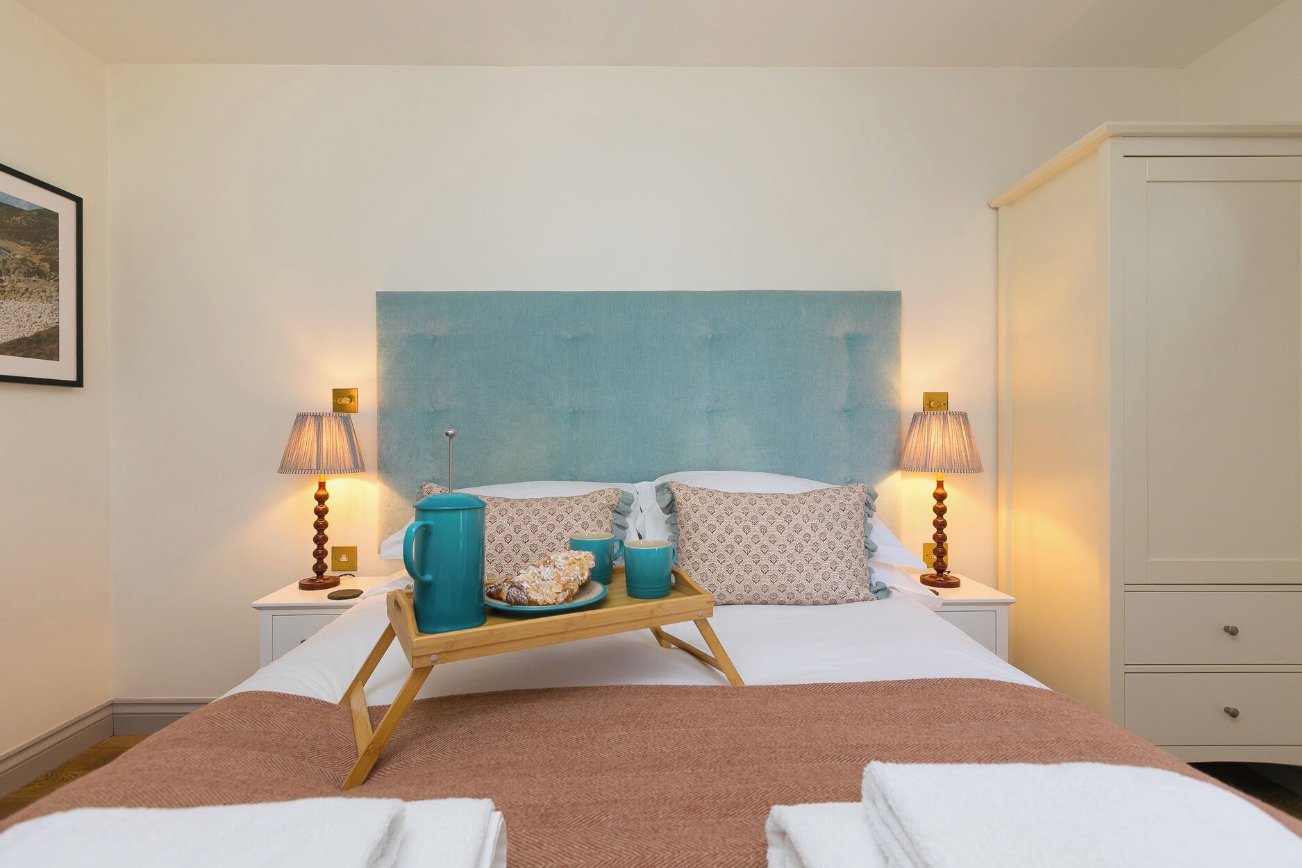 Une chambre d’hôtel avec un lit, deux tables de chevet, un plateau en bois avec des tasses et des pâtisseries, une grande tête de lit bleue et une armoire.