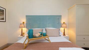 Une chambre d’hôtel avec un lit, deux tables de chevet, un plateau en bois avec des tasses et des pâtisseries, une grande tête de lit bleue et une armoire.